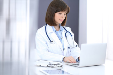 Doctor typing on laptop computer while sitting at the white table in hospital office. Physician at work. Medicine and healthcare concept