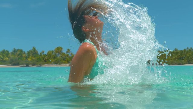 SLOW MOTION, CLOSE UP: Young woman pulls her head out of the glassy ocean and flips her hair back while cooling off by the tropical beach. Cheerful Caucasian girl playing in the turquoise exotic sea.