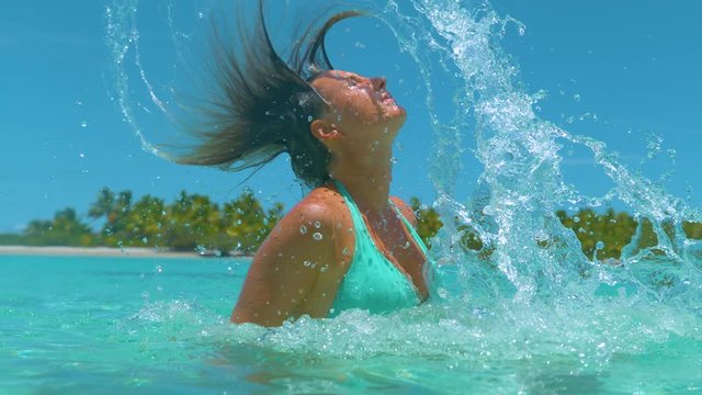 SLOW MOTION EFFECT, CLOSE UP: Young Caucasian woman pulls her out of the ocean and flips her long hair back, spraying glassy water everywhere. Cinematic shot of girl splashing water with her hair.