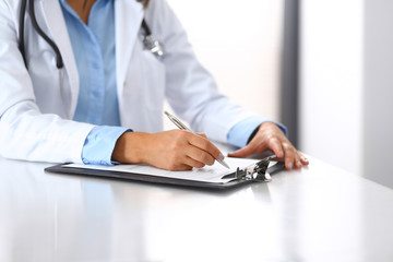 Unknown doctor woman filling up medical form while sitting at the desk in hospital office, close-up of hands. Physician at work. Medicine and health care concept