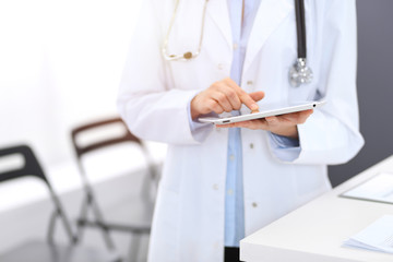 Female physician using digital tablet  while standing near reception desk at clinic or emergency hospital. Unknown doctor woman at work. Medicine concept