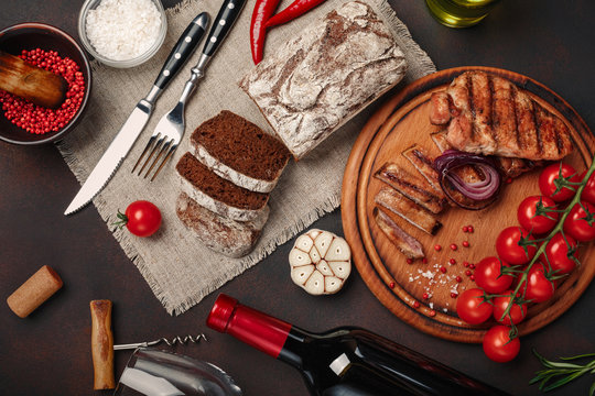 Sliced grilled pork steaks with bottle of wine, wine glass, corkscrew, knife, fork, black bread, cherry tomatoes, garlic, onion and rosemary on rusty background