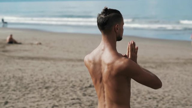 Backside view athletic shirtless man doing yoga on beach near ocean