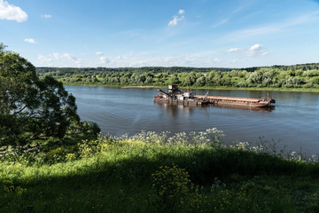Colorful landscape. Sand mining on the river. Special ship. Sunny summer day. Overgrown with wildflowers beach, tree. Far away on the horizon is a forest, a village.