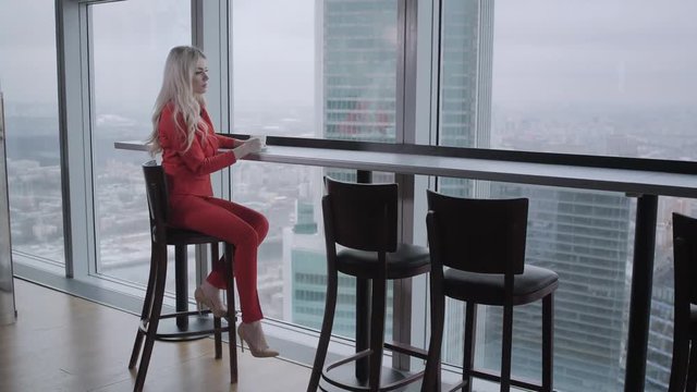 Woman in red coral business suit drinks coffee at a high table near the window. Young blonde businesswoman in the office in high floor in front of the window.