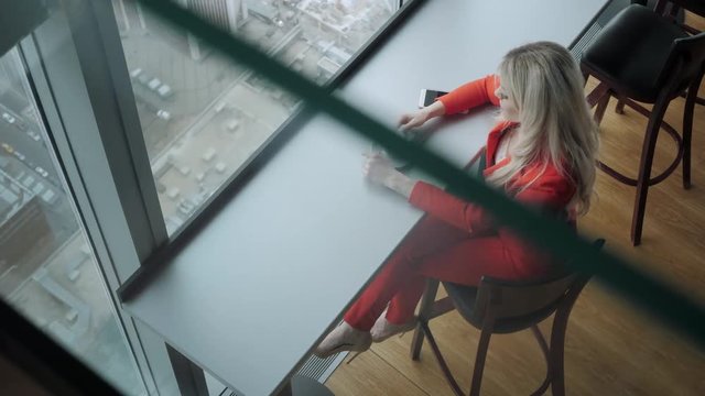 Woman in red coral business suit drinks coffee at a high table near the window. Young blonde businesswoman in the office in high floor in front of the window.