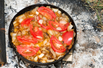 Tomatoes are fried on pork fat on a black pan on a metal grill during the journey for the preparation of scrambled eggs