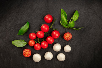 A flat lay composition with Mozzarella cheese, cherry tomatoes and fresh basil leaves on a black background, Italian cuisine ingredients with copy space