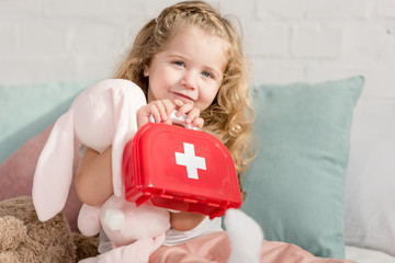adorable kid holding first aid kit and rabbit toy in children room, looking at camera
