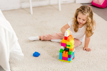 high angle view of adorable kid playing with colored plastic constructor on carpet in children room