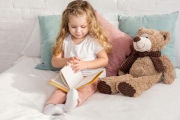 adorable child playing with teddy bear and reading book on bed in children room