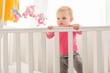 adorable toddler in pink shirt standing in crib and looking away