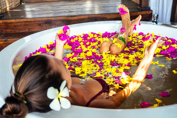 Back of woman in a bathtub with tropical exotic flowers in the water and flower white plumeria in her hair.girl in eco hotel taking bath in the spa salon summer vacation in a deluxe room ,skin care