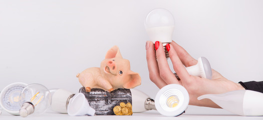 Female hand holds led lamp near the piggy bank with money on the background of lamps
