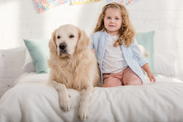 adorable kid and golden retriever dog sitting on bed in children room