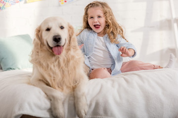 excited adorable kid and golden retriever sticking tongues out together on bed in children room