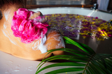Young beautiful woman relaxing in bath with foam and tropical flowers petals,tropical flowers petals in eco hotel Bali.brunette girl soaps the body with a sponge in jacuzzi exotic in paradise beach