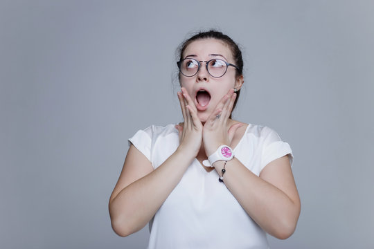 The girl shocked by something  what she saw. Young woman in glasses and in white blouse standing on th background in studio and posing on the camera. She open her mouth and pressed her hand to cheecks