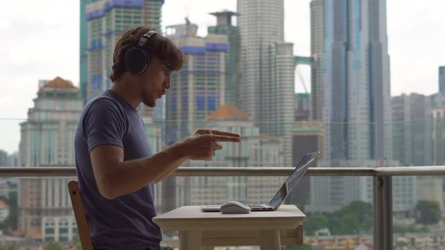 Young man freelancer workes on his laptop at a balcony with a background of a city center full of skyscrapers. Remote techer has a conversation with a student through the internet