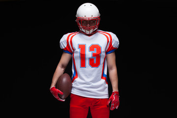 American football player wearing helmet posing with ball on black background