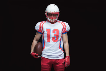 American football player wearing helmet posing with ball on black background