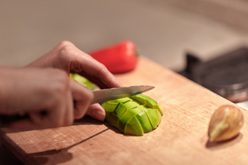 Female hands cutting fresh organic avocado with knife on wooden board in kitchen. Red bell pepper on background. Close up, indoors, copy space.
