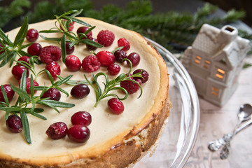 Festive cheese cake with raspberries, rosemary and lingonberries on the background of a fir branch and a toy house