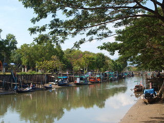Fototapeta na wymiar Traditional fishing Thai boats on the river. Channel with muddy water and old Asian fishing boats moored at the edge. Longtailboats in Thailand. Longtail wooden boat, water and green plants and trees
