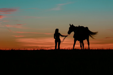 Graceful girl walking with horse and holding reins in hand. Romantic equine and girls silhouette on horse hiking with red rising sun on horizon