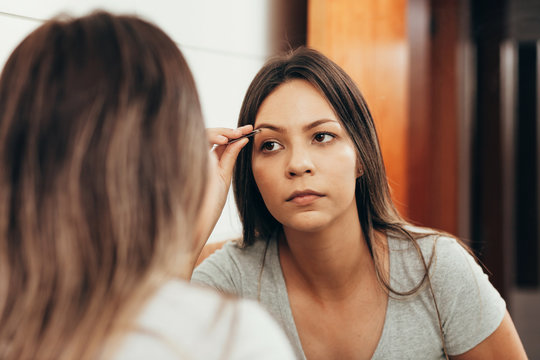 Young woman plucking eyebrow with tweezers in front of mirror