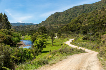 Fototapeta na wymiar Dirt road with a river and forest beside and mountains in the background, blue sky with few clouds, Urubici, Santa Catarina