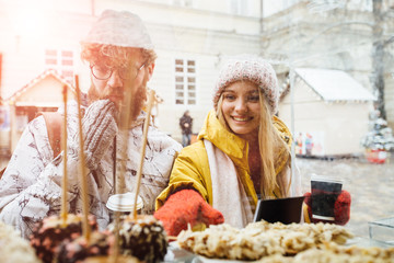 Excited hipster couple choosing cake while spending time together in outdoors winter christmas fair. Surprised happy blond woman and beard caucasian man point in show case by hand in red mitten.