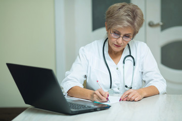 stylish aged woman in a white coat sits at the table in the hospital with a laptop writes documents