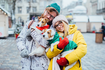 Lovely family portrait: funny hipster beard man and blond woman both in eyeglasses hugging their two Jack russel terrier outdoor in winter city on background. Dogs wearing warm red and green jacket.