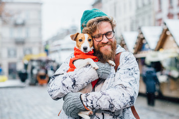 Portrait of funny beard hipster caucasian man closed eyes in eyeglasses embracing his Jack russel terrier dog in red jacket, looking at camera and smiling in outdoor winter market square european city
