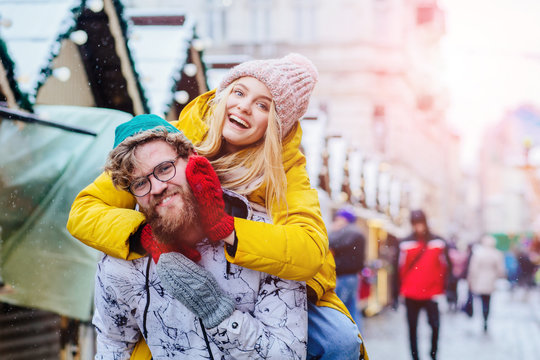 Image of beard caucasian man having fun and giving piggyback ride to joyful blond woman during winter city street. Sun glare effect. Sparkling with happiness concept