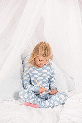 Fototapeta premium cute kid in pajamas sitting on bed with crossed legs and writing in notebook