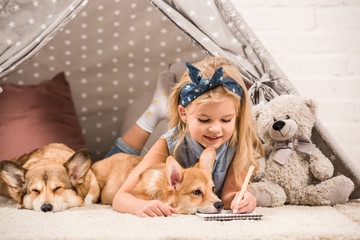 adorable child lying in wigwam with welsh corgi dogs and writing in notebook at home © LIGHTFIELD STUDIOS