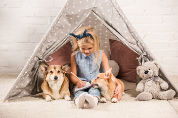 cute child sitting with corgi dogs in wigwam and writing in notebook at home © LIGHTFIELD STUDIOS