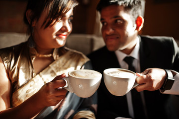 Elegant and fashionable indian friends couple of woman in saree and man in suit sitting on cafe and drinking cappuccino.
