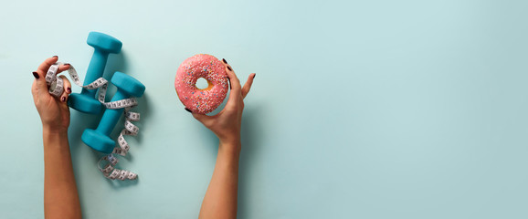 Female hand holding sweet donut, measuring tape, dumbbells over blue background. Top view, flat...