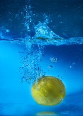 Lemon falls into water with splashes and bubbles, on blue background.