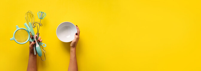 Woman hand holding kitchen utensils on yellow background. Baking tools - bowl, brush, whisk,...