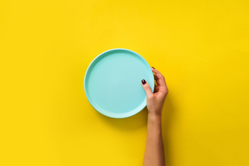 Female hand holding empty blue plate on yellow background with copy space. Healthy eating, dieting concept. Banner