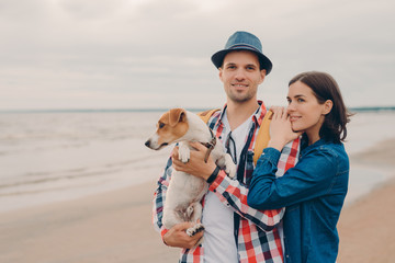 Outdoor shot of pleased man and woman stand closely with favourite dog, look in distance, enjoy...