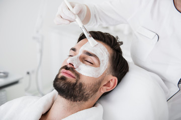 Cosmetologist using brush while putting white clay mask on male face