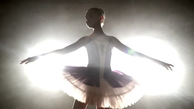 Ballerina's silhouette turning around. ballerina with hair in a bun is dancing on a scene, against bright light of soffit and smokescreen
