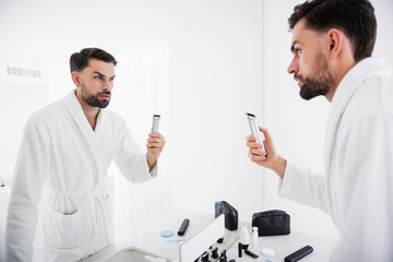 Waist up of serious man standing with his shaving machine in the bathroom