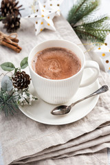 Obraz na płótnie Canvas Cup of hot chocolate with cinnamon sticks on the white wooden table. Winter cocoa drink on a napkin, Christmas-tree golden stars decorations, branches and cones