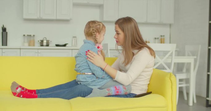 Caring affectionate mother comforting her upset adorable daughter while sitting face to face on cozy sofa in domestic room. Loving single mom consoling sad pensive elementary age girl at home.